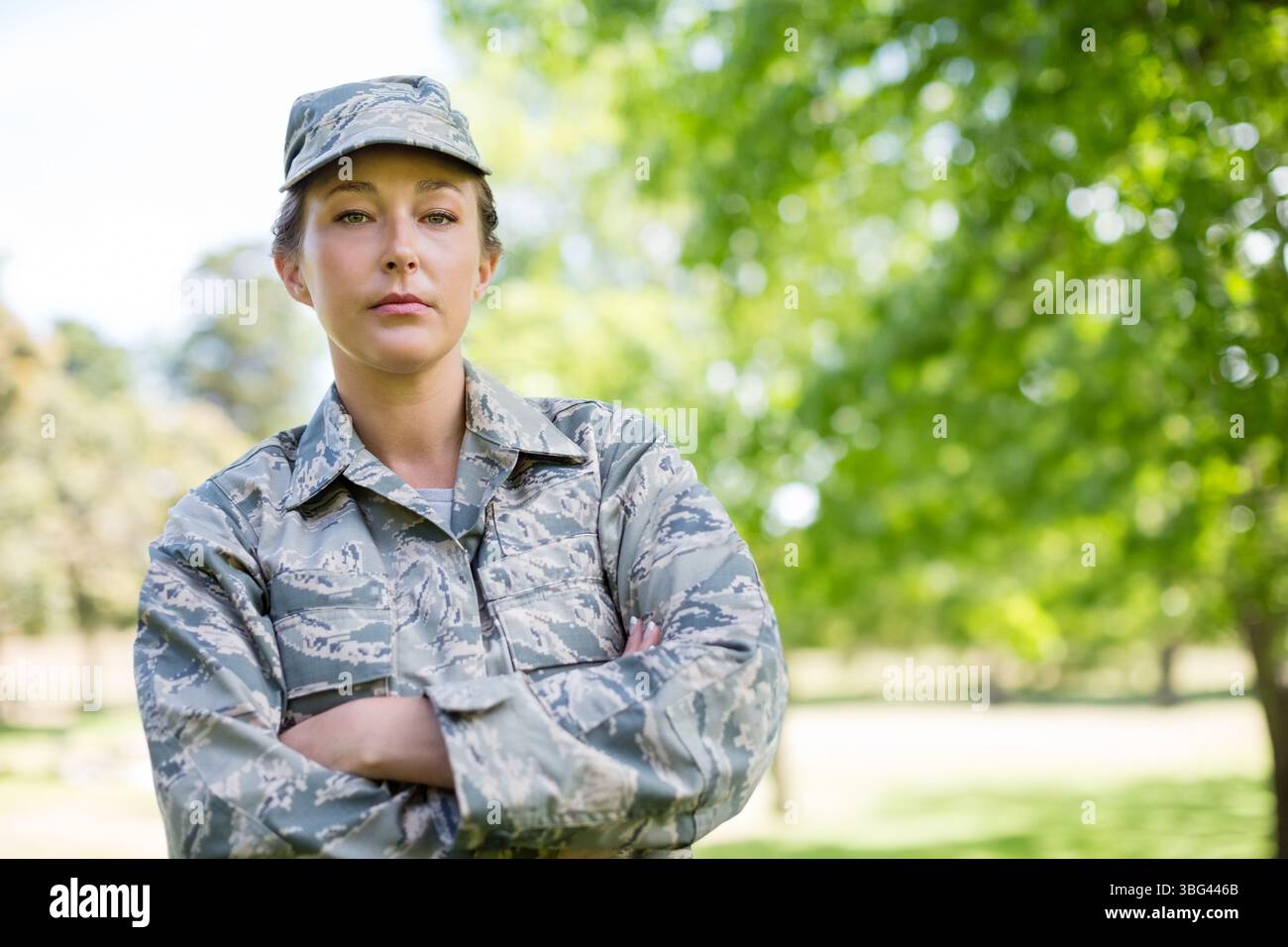 Mid adult female soldier standing with arms crossed in grassy park in camouflage uniform Stock Photo
