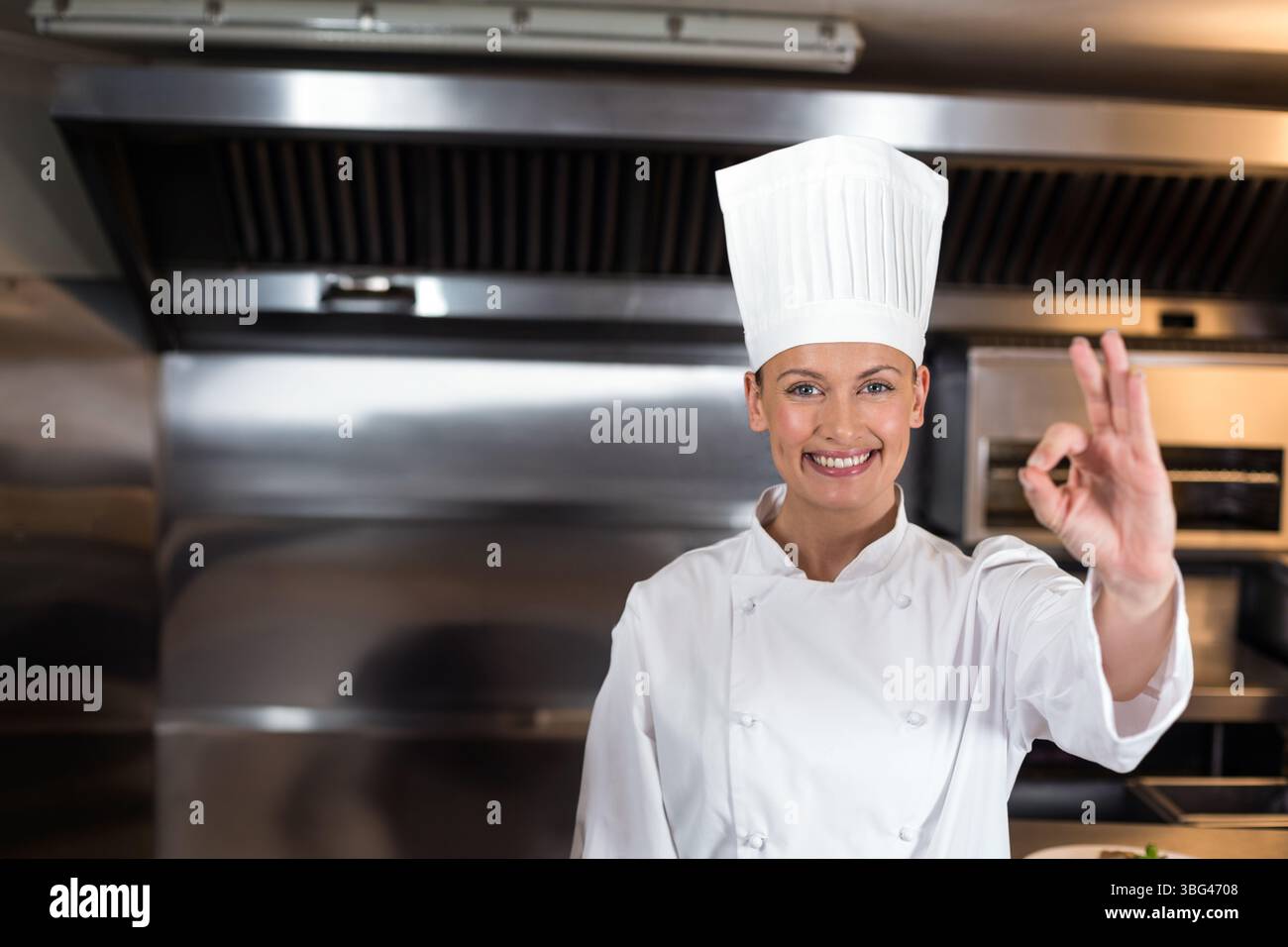 Female chef wearing chef coat and toque standing under hood beside oven ...