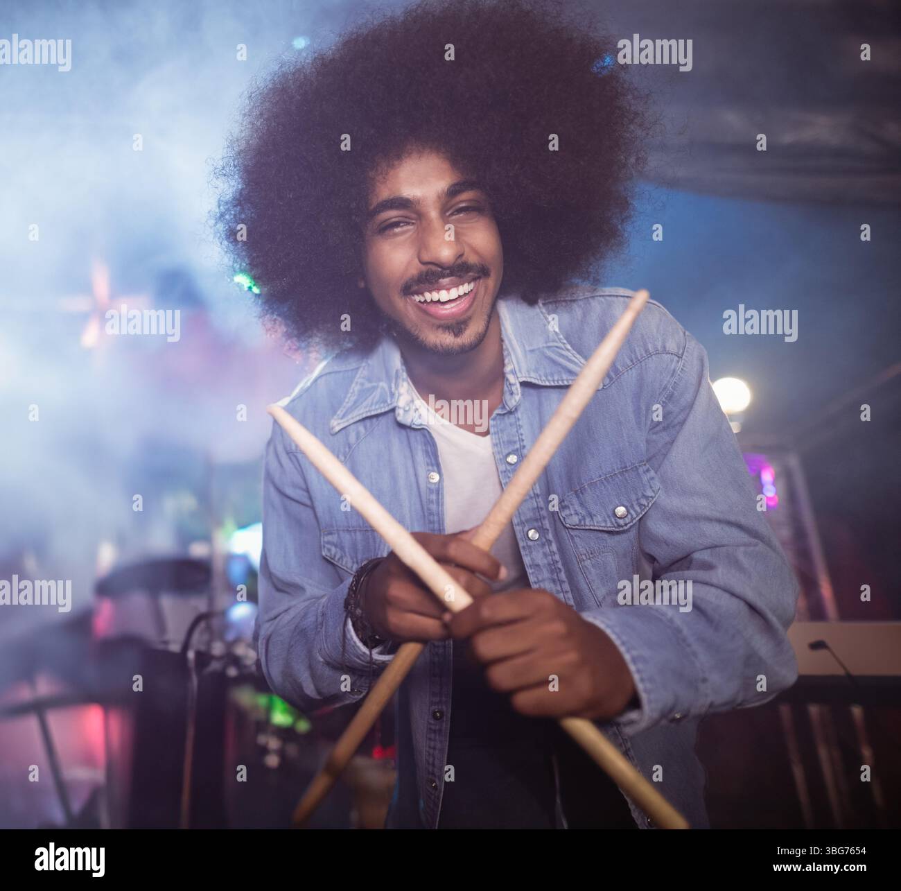 Indian man standing on concert stage holding wooden drumsticks amid colored lights and fog Stock Photo