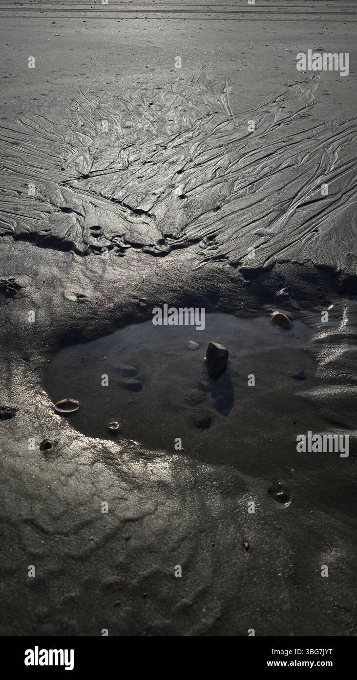 The tide recedes, leaving a pristine small pool of water on the sandy beach, reflecting the sky. In the exact center and a smooth stone stands out - Smartphone Captured Stock Image