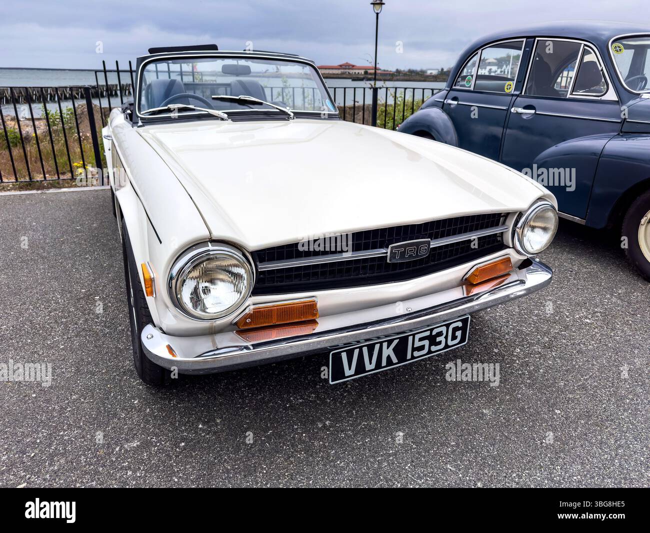 Front view of a classic white TR6 convertible car parked on a paved area beside an iconic vintage car under a cloudy sky - Smartphone Captured Stock Image
