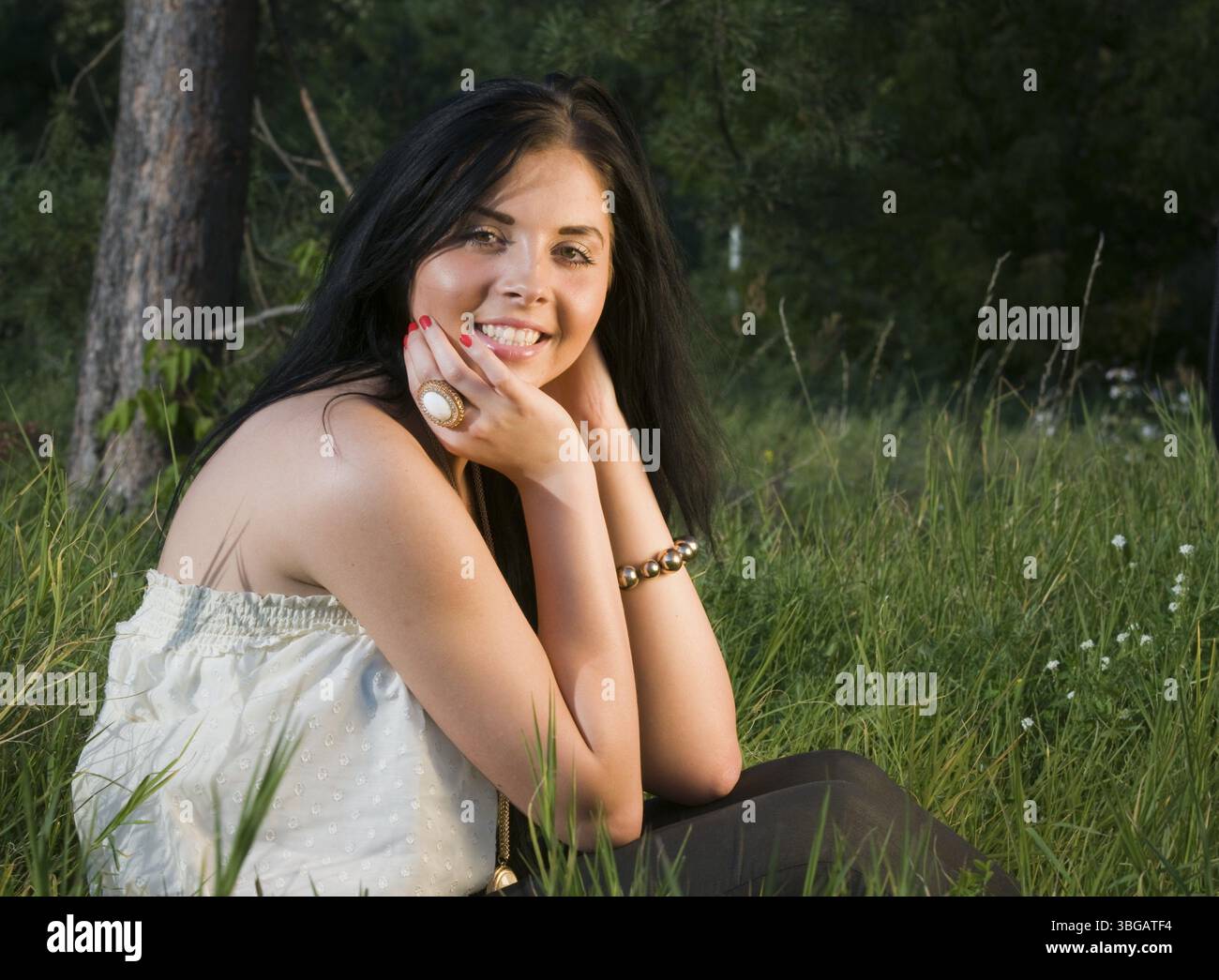 Young black-haired woman sitting in the grass resting her elbows on her knees and smiling into the camera Stock Photo