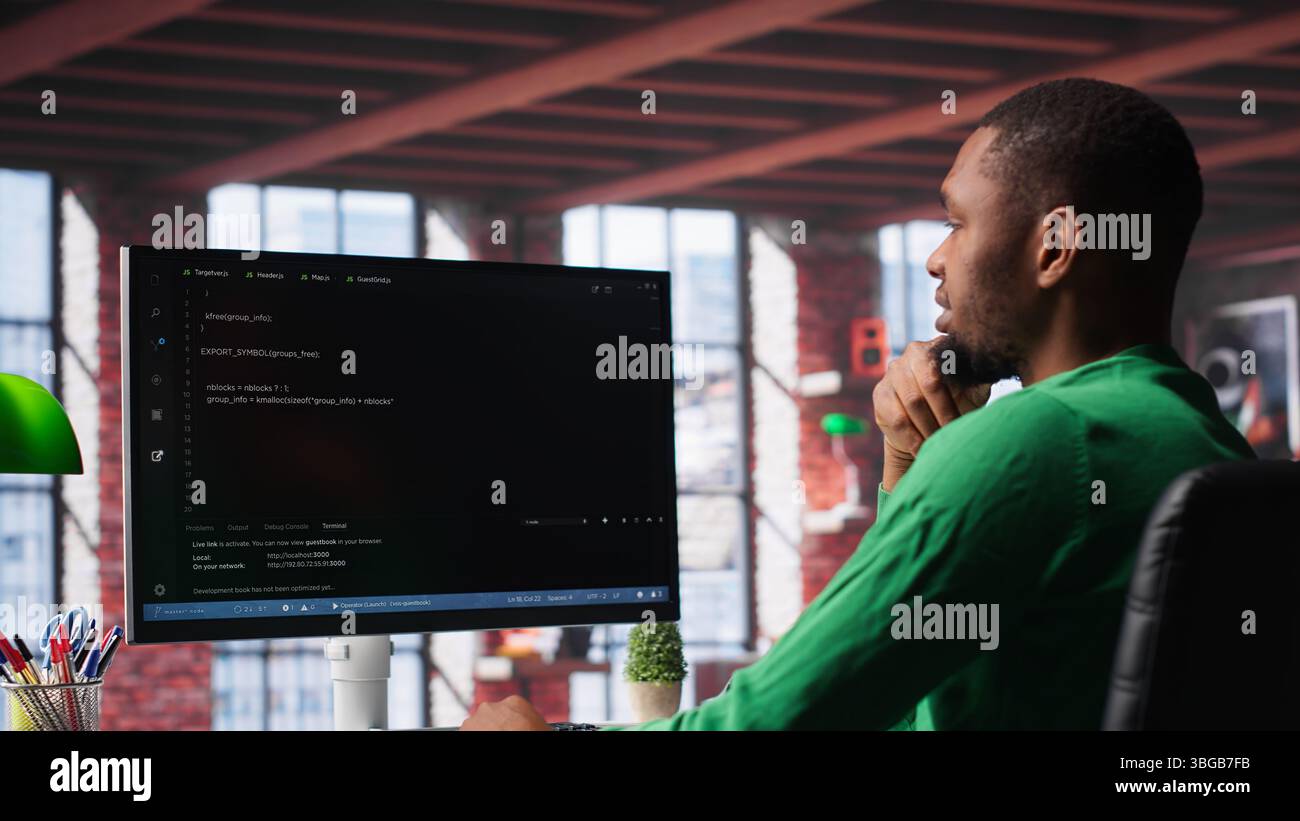 Black technician in home office looks at UI on computer monitor, typing and executing programming code. IT worker using coding application to write and test code in modern apartment. Camera A. Stock Photo