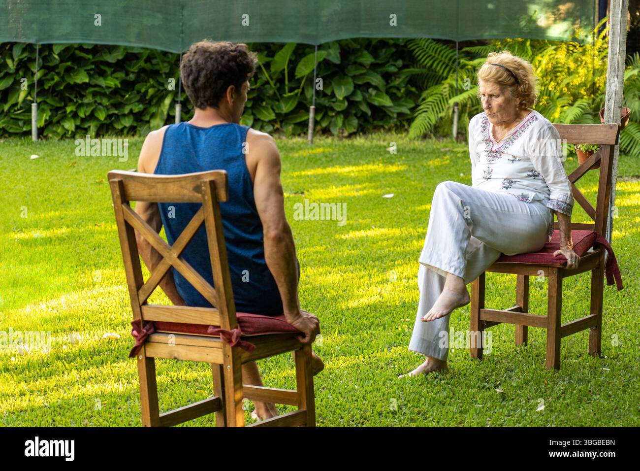 Senior woman and her physiotherapist are sitting on chairs in a garden, practicing rehabilitation exercises to improve mobility and well-being Stock Photo