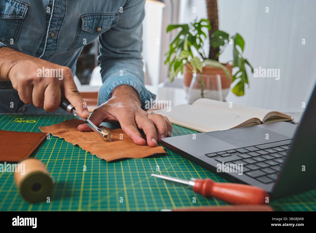 Craftsman working leather using stitching wheel in his workshop with laptop, notebook and tools Stock Photo