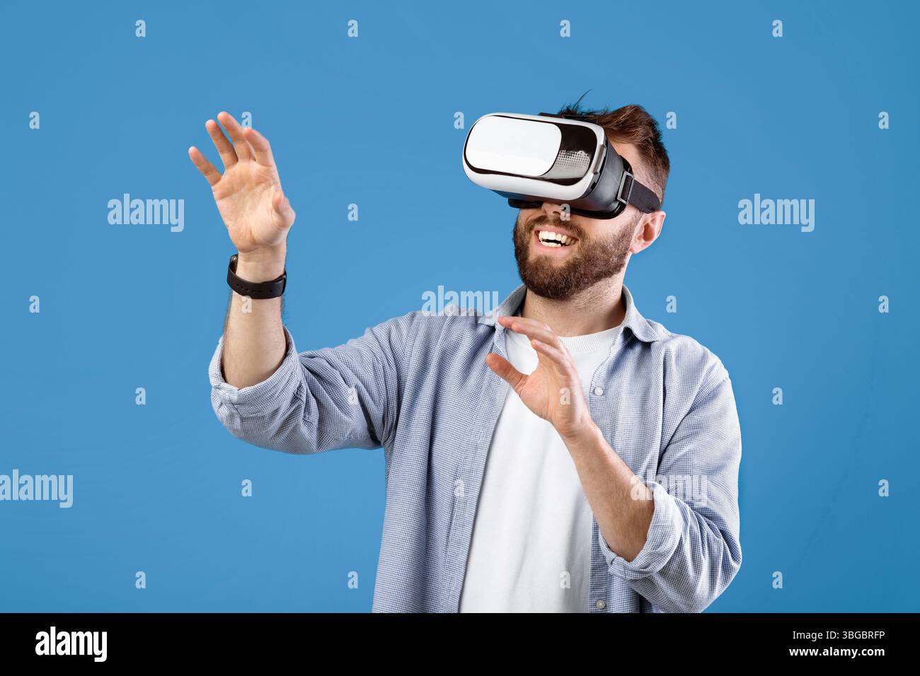 Happy bearded young man using VR headset to explore augmented reality on blue studio background Stock Photo
