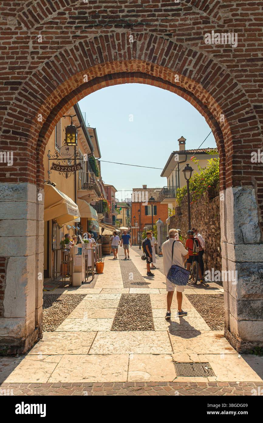 Garda Lake, Italy 3 January 2025: Tourists walking in a picturesque street in lazise city center under a brick arch near garda lake, italy Stock Photo