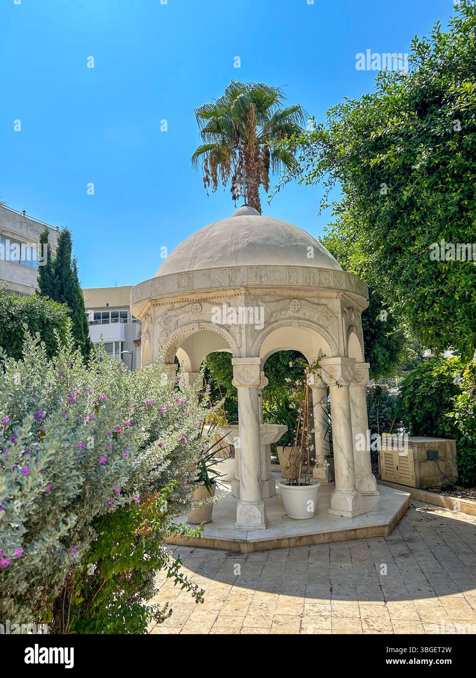 A decorative marble gazebo with arches and columns surrounded by lush greenery and potted plants on a sunny day in a Mediterranean-style courtyard. - Smartphone Captured Stock Image