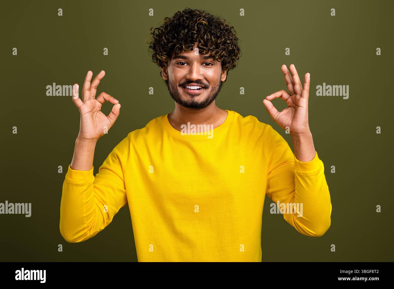 Young curly haired man in yellow shirt making OK gesture with both hands against khaki background Stock Photo