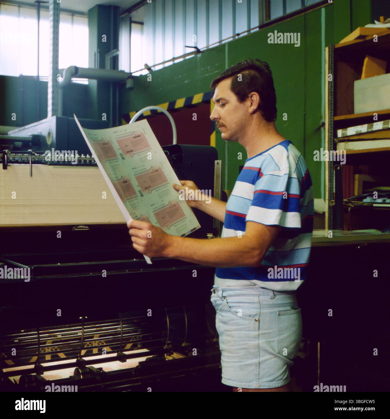 Ein Angestellter der Papierwerke Waldhof-Aschaffenburg (PWA) steht vor einer Druckmaschine und prüft einen frisch bedruckten Bogen. Die Druckbögen zeigen Etiketten oder Verpackungselemente. Der Arbeitsplatz befindet sich in einer Industriehalle mit grün gestrichenen Wänden und Regalen voller Materialien im Hintergrund. Das Bild entstand wahrscheinlich in der Firmenzentrale in Raubling bei Rosenheim, circa 1983. Stock Photo