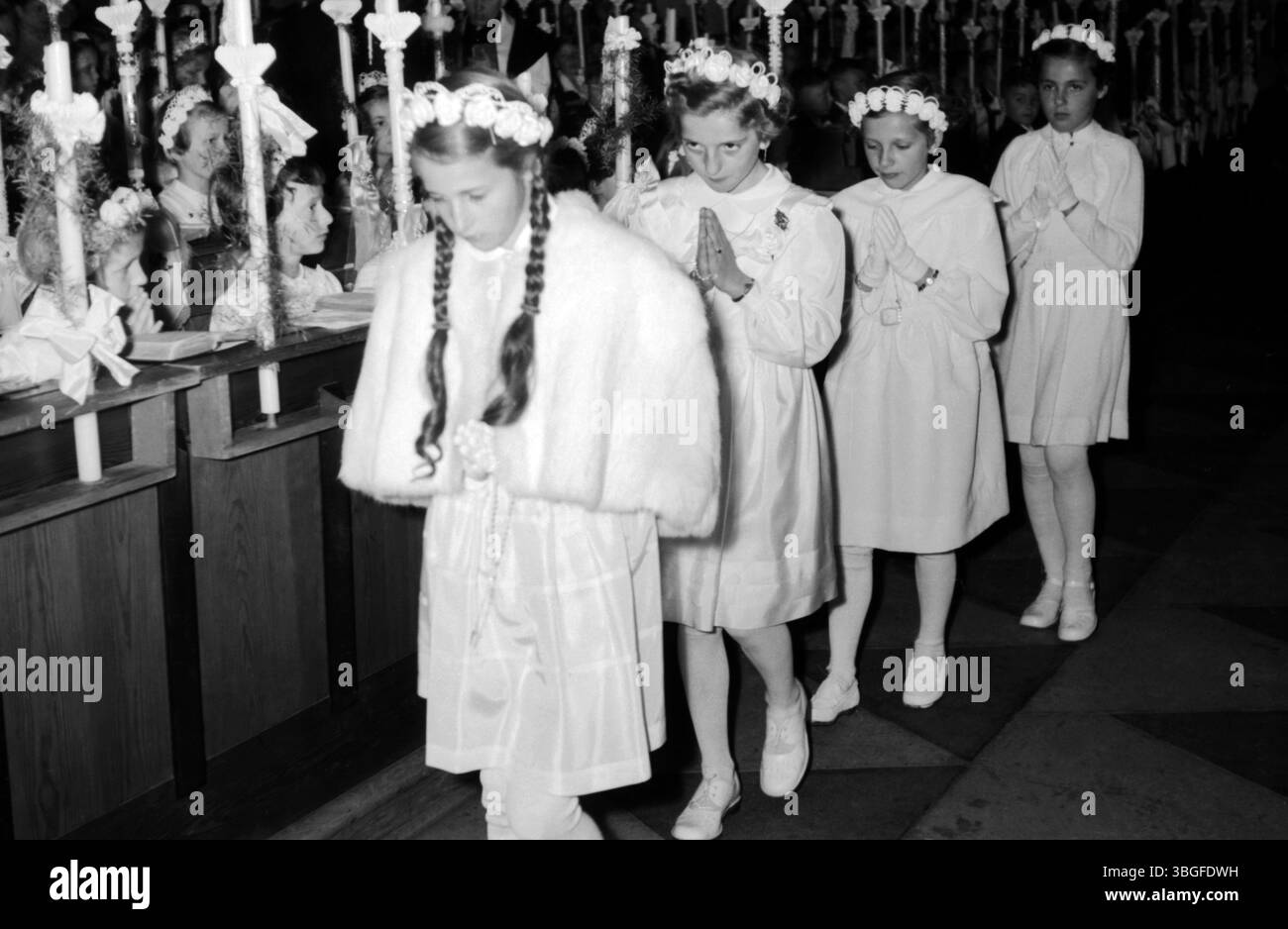 Mädchen in weißen Kleidern und Blumenkränzen ziehen betend durch den Mittelgang einer Kirche. Die Szene zeigt eine feierliche Erstkommunion, Anfang der 1950er Jahre, festlich geschmückte Kerzen stehen an den Kirchenbänken. Stock Photo