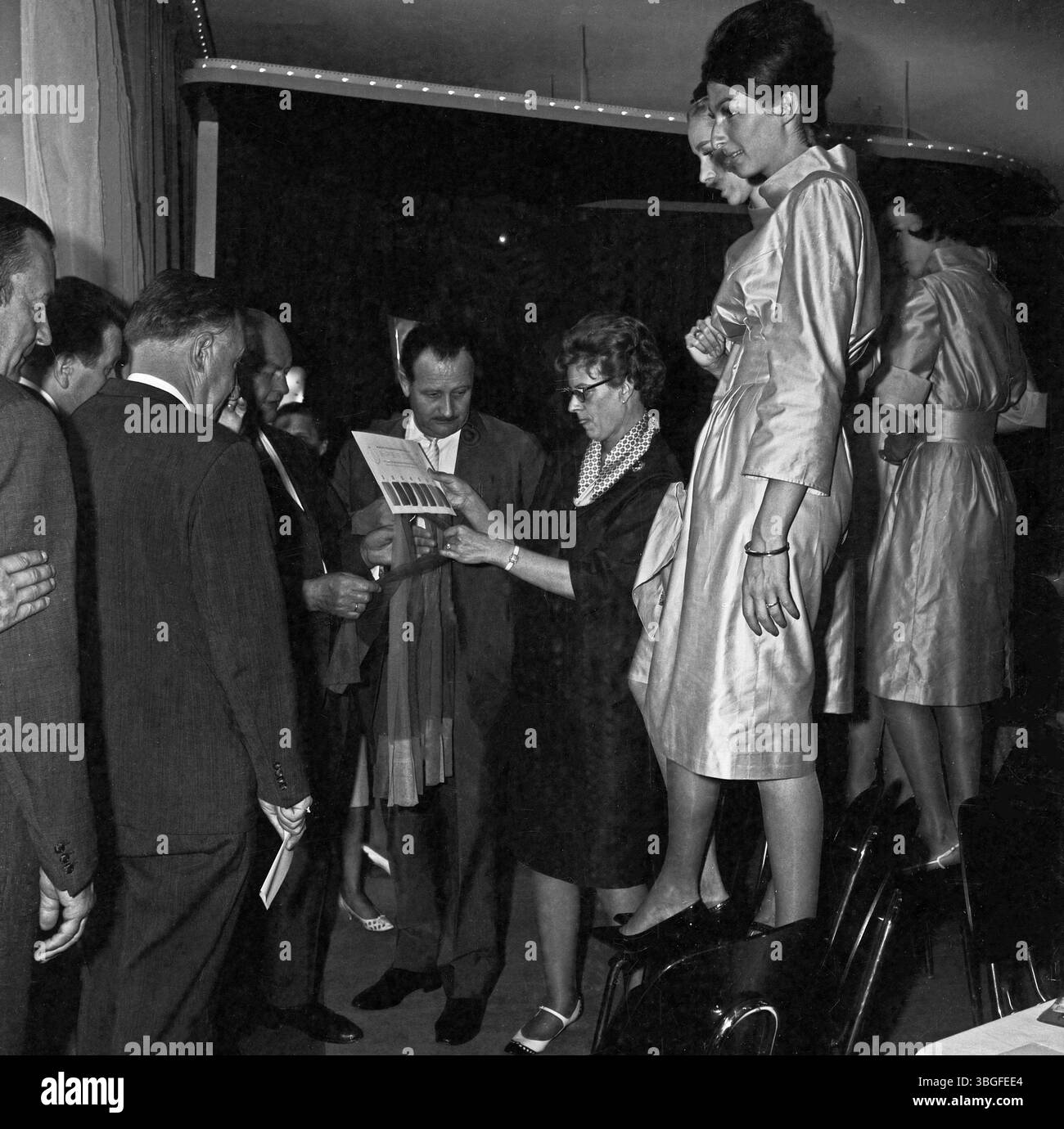 Bei einer Hausmesse für Damenstrumpfhosen, ca. 1962, besprechen Einkäufer und Vertreter die angebotenen Modelle direkt vor den präsentierenden Mannequins. Eine Verkäuferin zeigt Farb- und Materialmuster, während die Models auf Stühlen stehend die Strümpfe am Bein tragen. Stock Photo