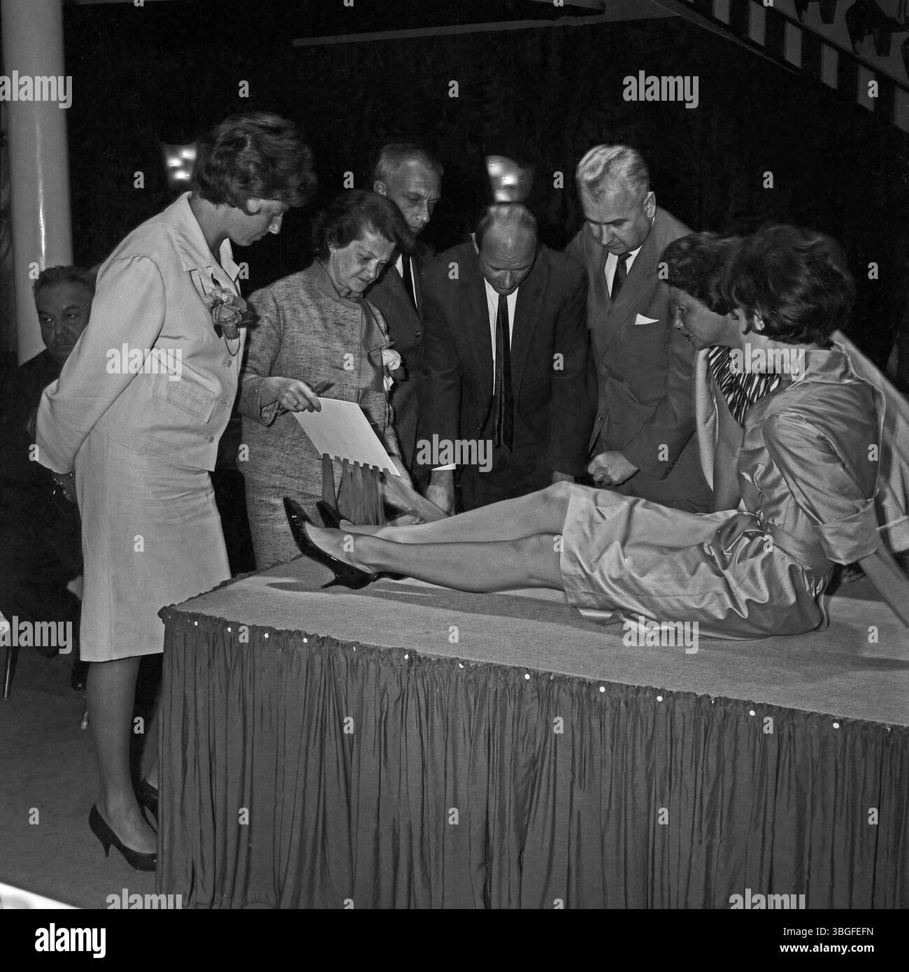 Auf einer Hausmesse für Damenstrumpfhosen um 1962 begutachten Einkäufer direkt am Podest die Qualität der präsentierten Strümpfe, die ein Model im Sitzen trägt. Die direkte Nähe zwischen Produkt und Publikum zeigt den praktischen, verkaufsorientierten Charakter der Veranstaltung. Stock Photo