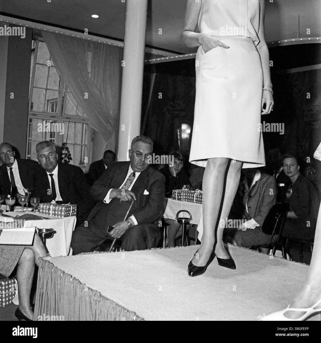 Eine Frau präsentiert auf einem Laufsteg ein ärmelloses Etuikleid während einer Hausmesse für Damenstrumpfhosen um 1962. Die Blicke der männlichen Einkäufer sind auf das Modell gerichtet, während im Hintergrund weitere Gäste aufmerksam das Geschehen verfolgen. Stock Photo