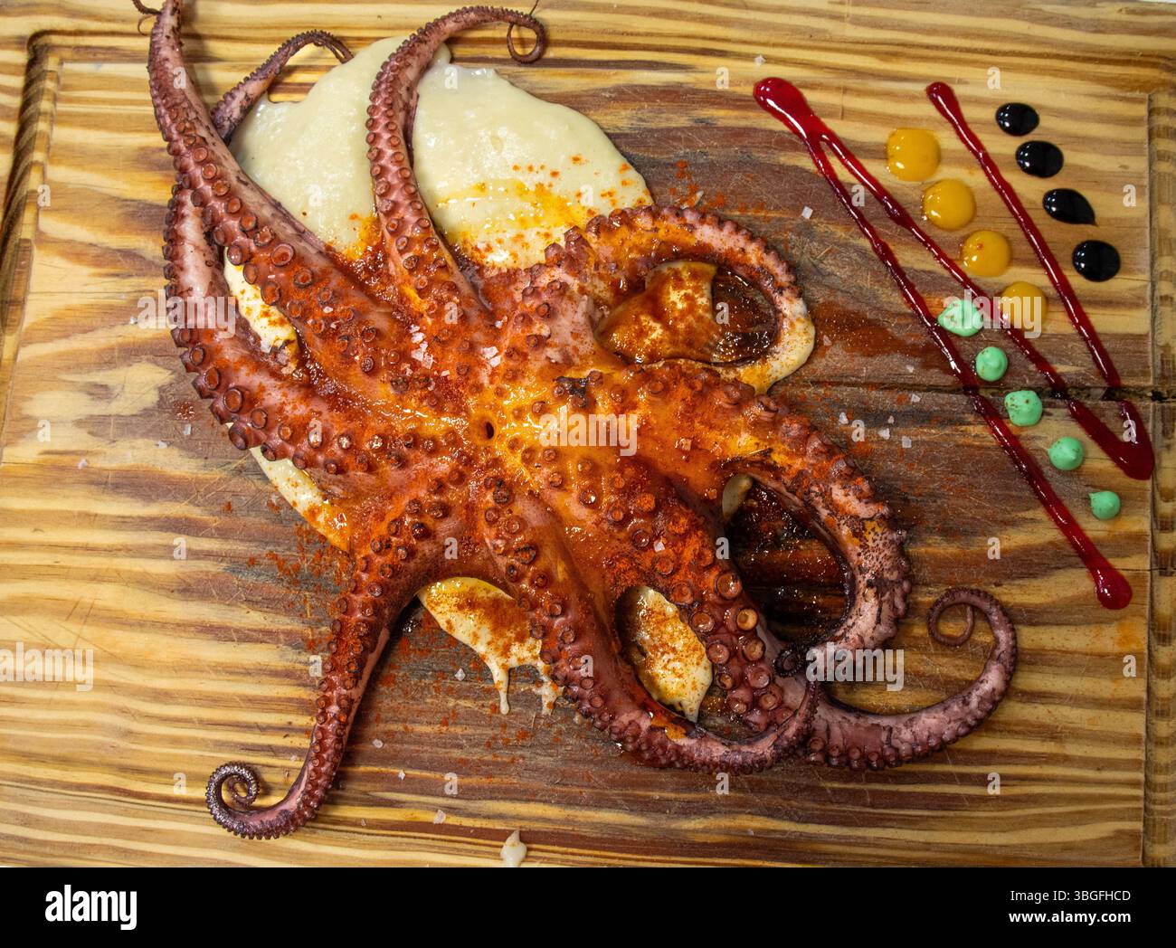 Pulpo asado presentado sobre una tabla de madera rústica. Condimentado con pimentón y aceite de oliva y a un lado,líneas y puntos de salsas de colores. Stock Photo