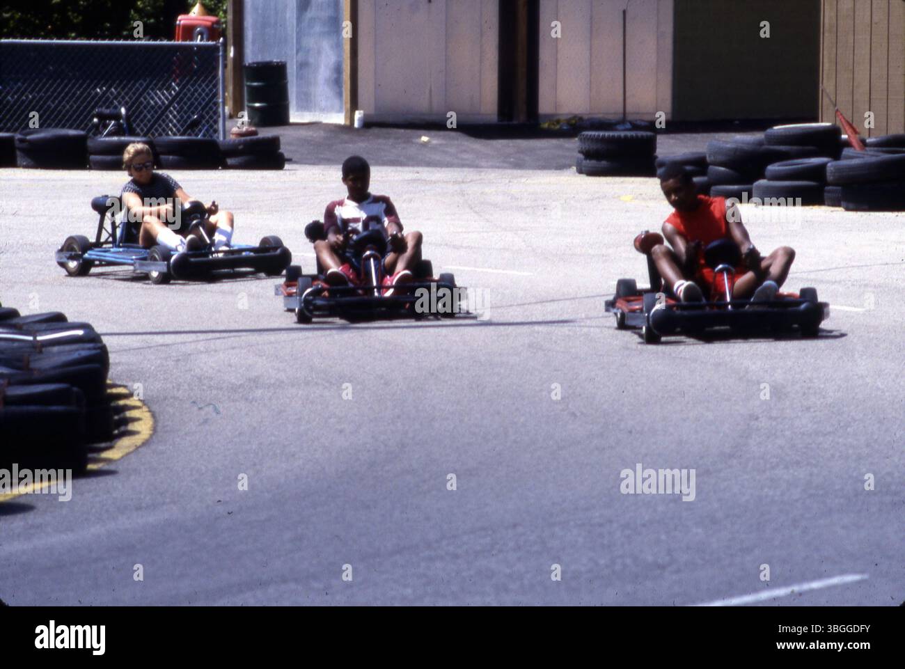 Three children drive mini race cars around a track at the Midge-o-racer attraction in Kiddie Land, Wyandot Lake Amusement Park, August 1985. A red and white gas pump stands behind a fence. Stock Photo