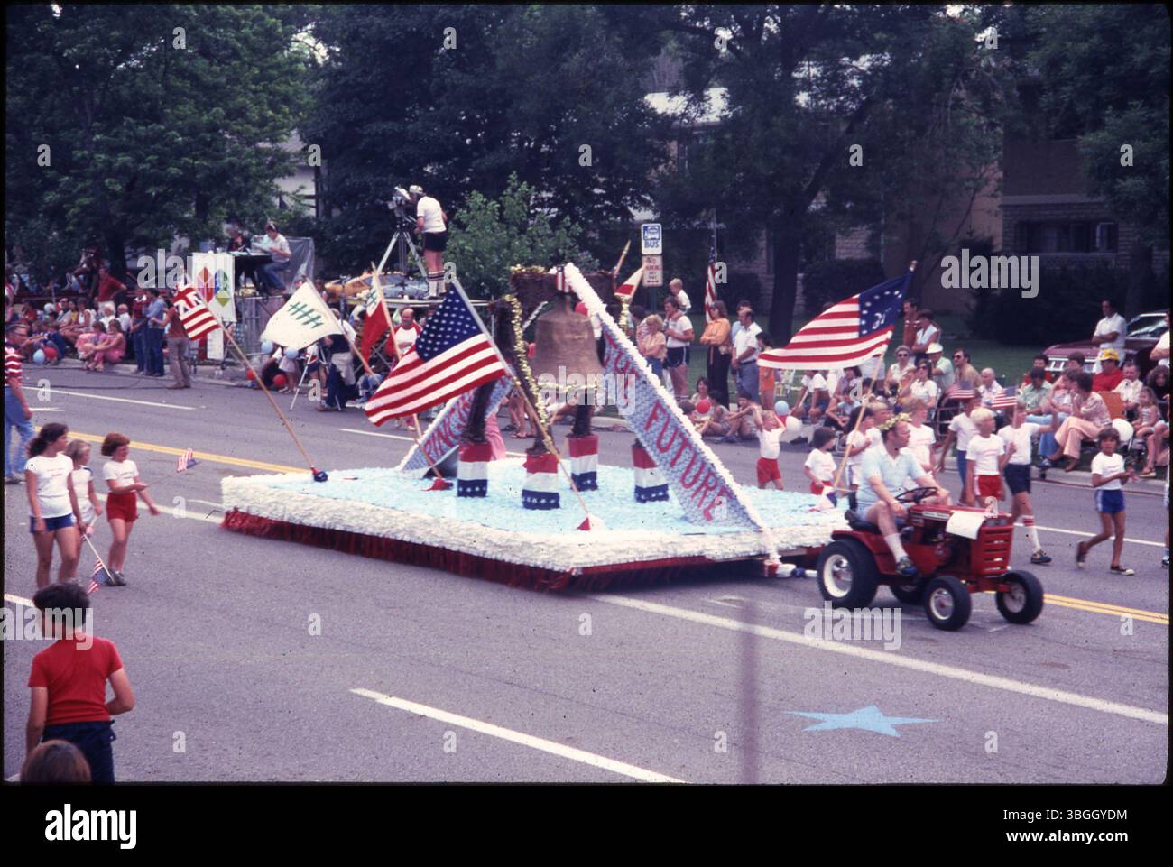 The 1980 Upper Arlington Fourth of July Parade featured a red, white, and blue float with a replica of the Liberty Bell and the banner 'Ringing in the Future.' The float was pulled by a red lawn tractor and accompanied by children wearing U.S. flag-themed outfits. Stock Photo