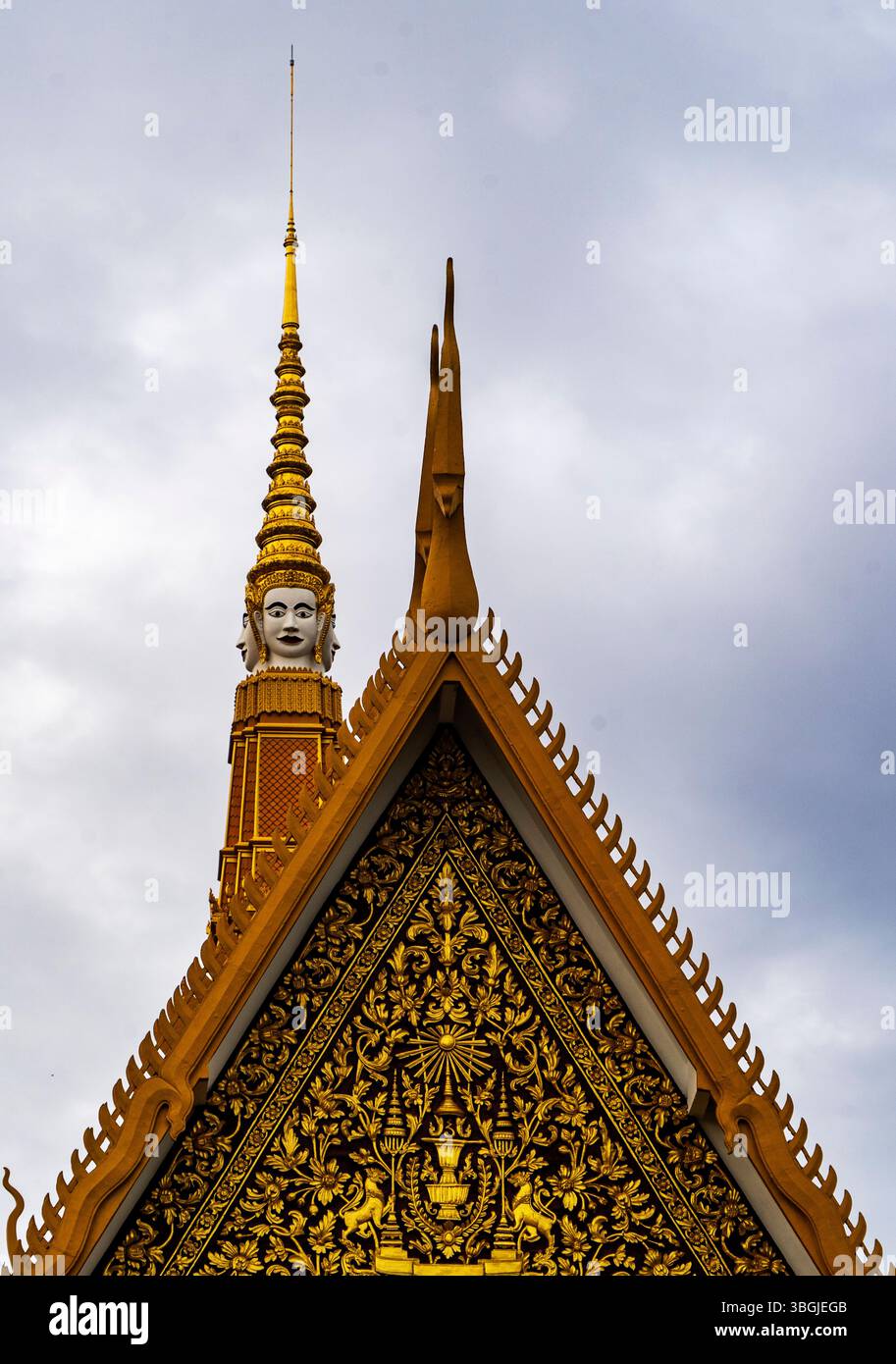 A close-up view of a traditional Thai temple roof featuring intricate golden decorations and a tall spire topped with a statue. The sky is overcast, a Stock Photo