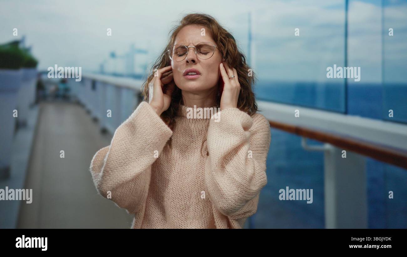 Woman on cruise ship with eyes closed and hands on head, appears seasick, wearing sweater, situated outdoors on the deck overlooking the sea, showing Stock Photo