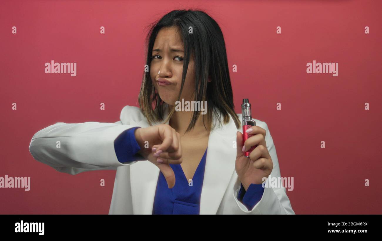 Woman expressing disapproval while holding an e-cigarette against a vibrant  red background, demonstrating a negative reaction to vaping Stock Photo