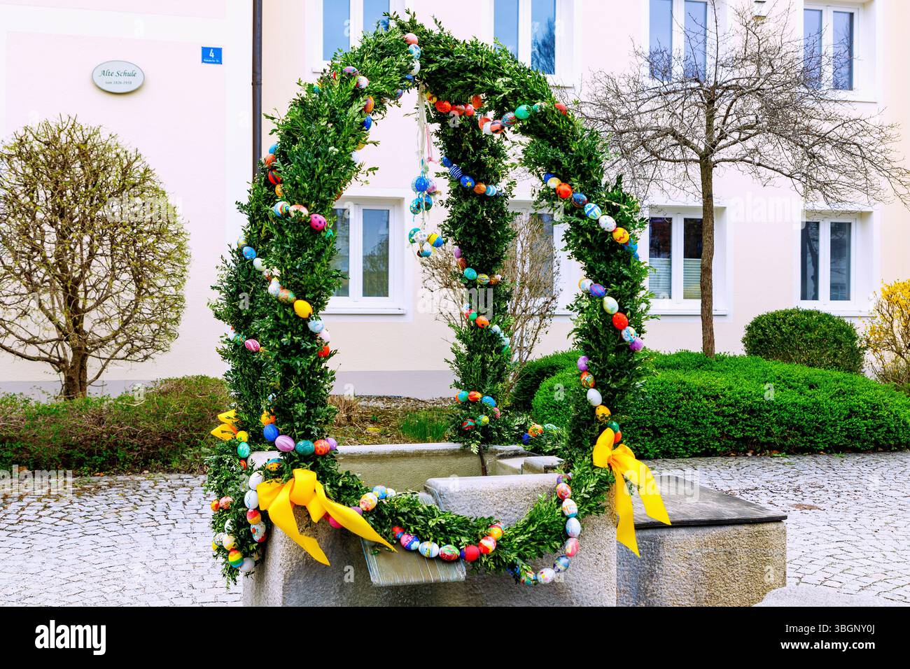 Easter fountain decorated with garlands of beech branches and colorful Easter eggs in front of the old school in Forstinning in Upper Bavaria in Germany Stock Photo