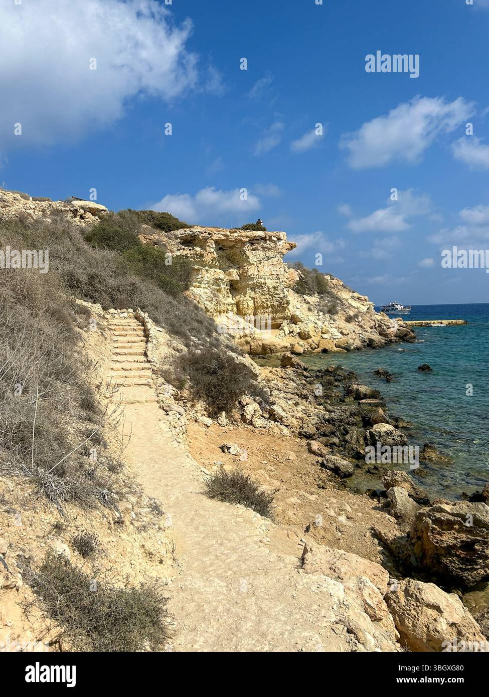 Scenic coastal trail with stone steps winding through rocky cliffs and dry vegetation, overlooking the clear blue sea under a bright sky. - Smartphone Captured Stock Image