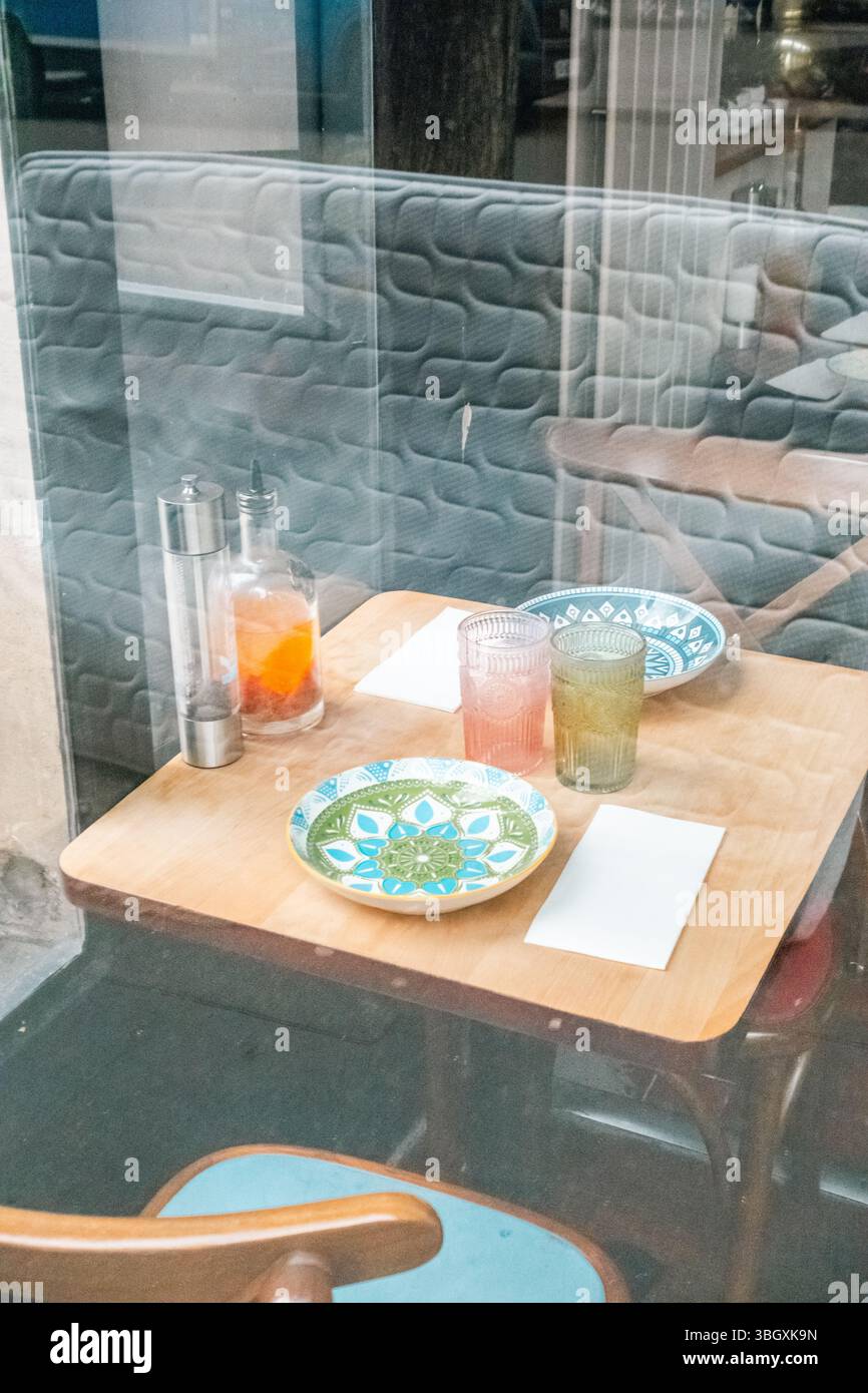 A view through a cafe window showing a set wooden table with colorful patterned plates, drinking glasses, and napkins. Reflections are visible on the Stock Photo