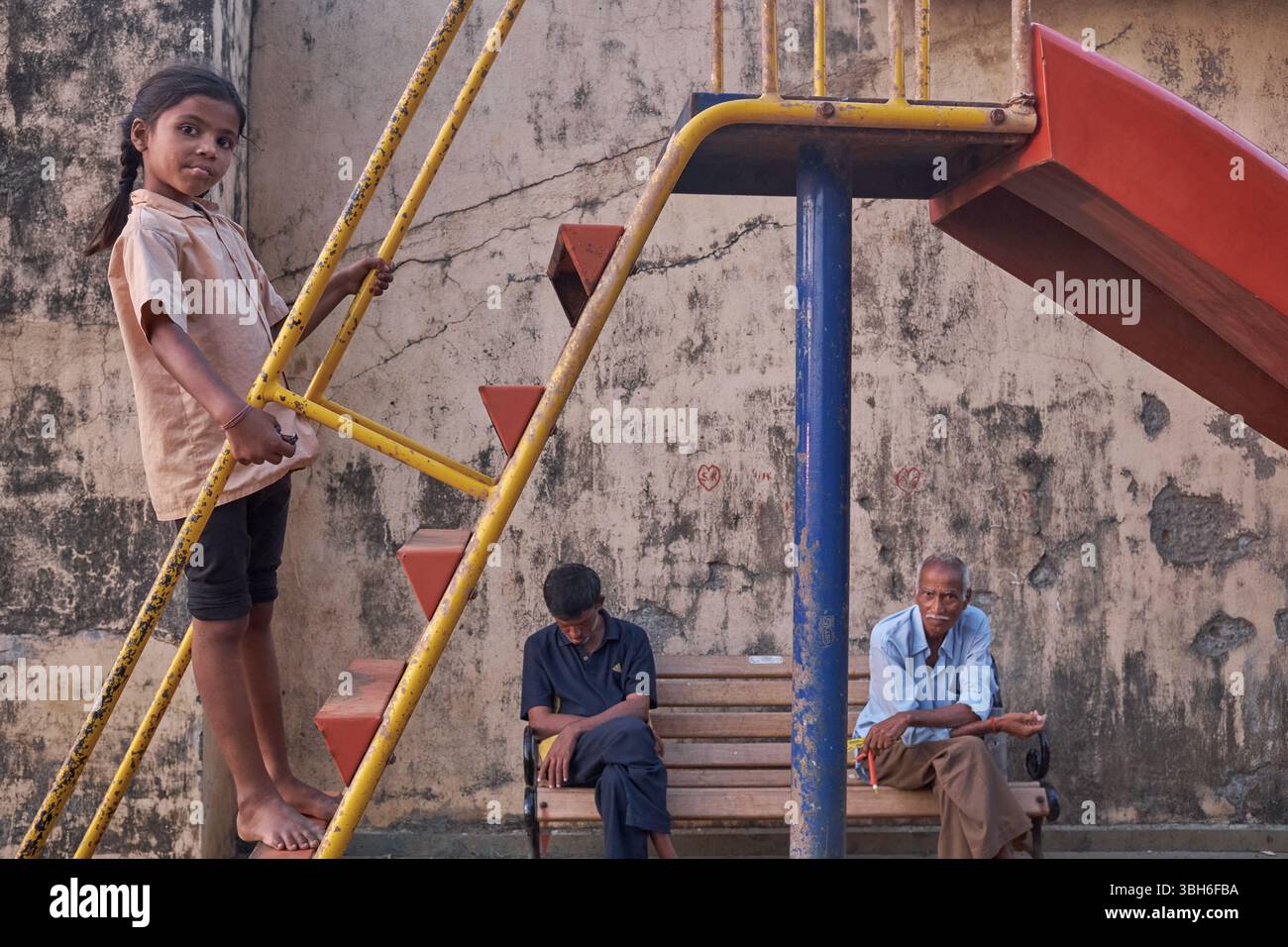 An Indian girl child stands on the ladder of a playground slide, looking cat the camera, two grown-ups in the background; Mumbai, India Stock Photo
