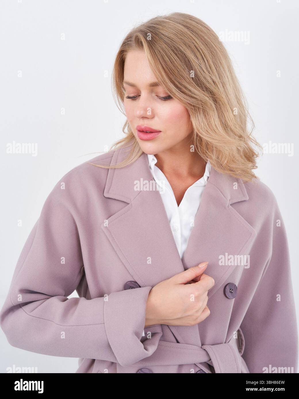 Blonde woman wearing a light mauve trench coat over a white blouse, with her eyes gently closed, exuding a serene demeanor and softly styled loose Stock Photo
