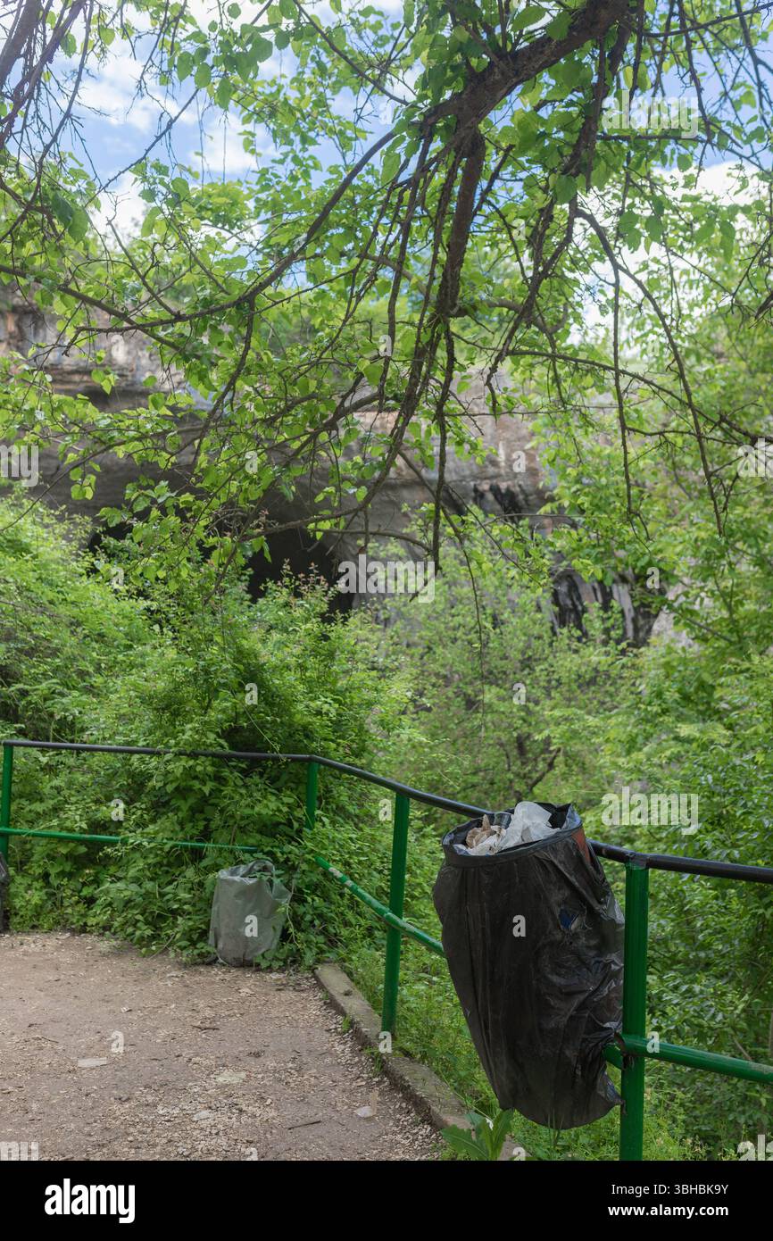 Overflowing plastic garbage bag tied to green rail in lush forest path. Stock Photo