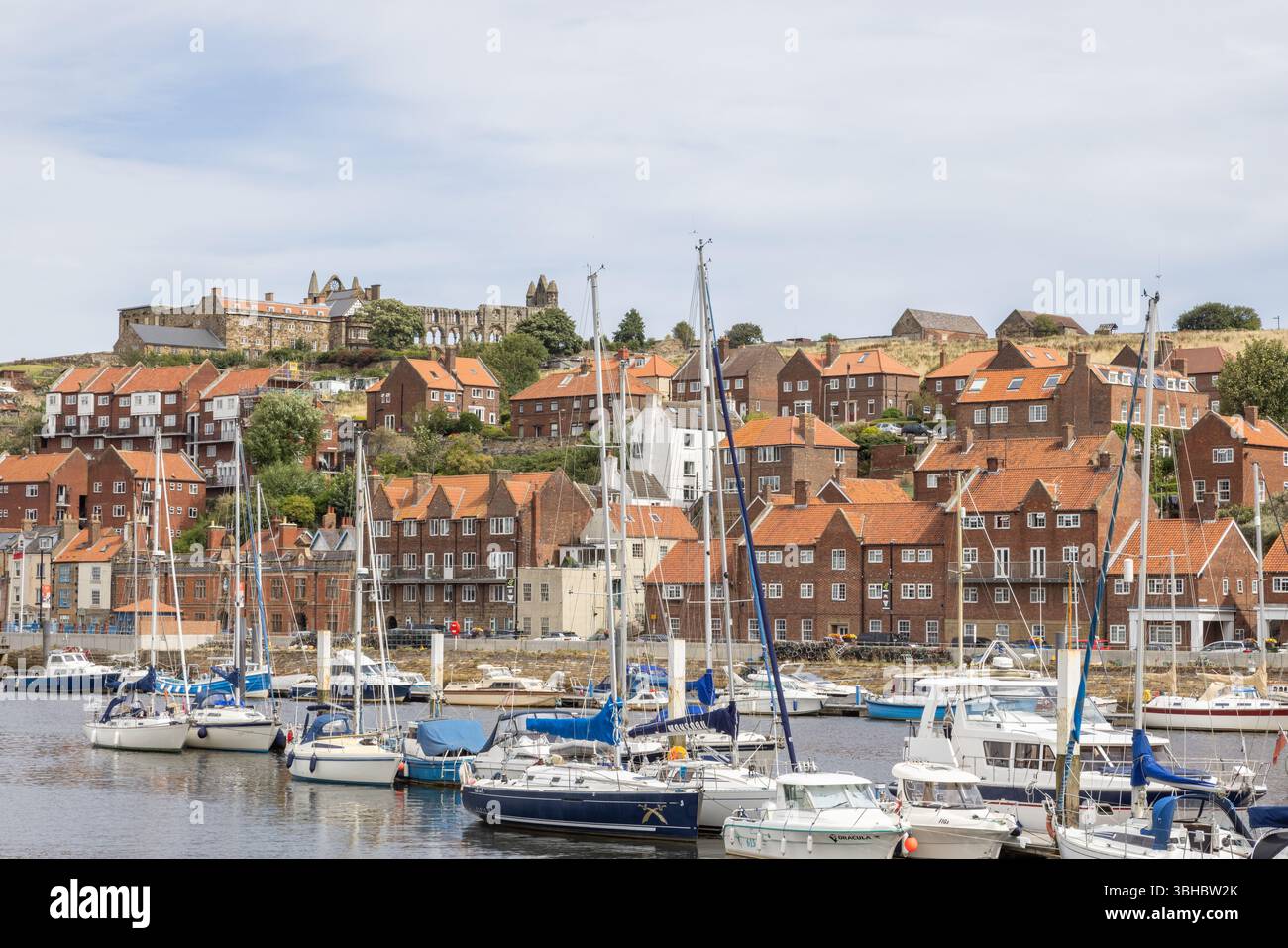 Whitby harbour with yachts moored along the River Esk, red roofed cottages and the ruins of Whitby Abbey on East Cliff in the background. Stock Photo