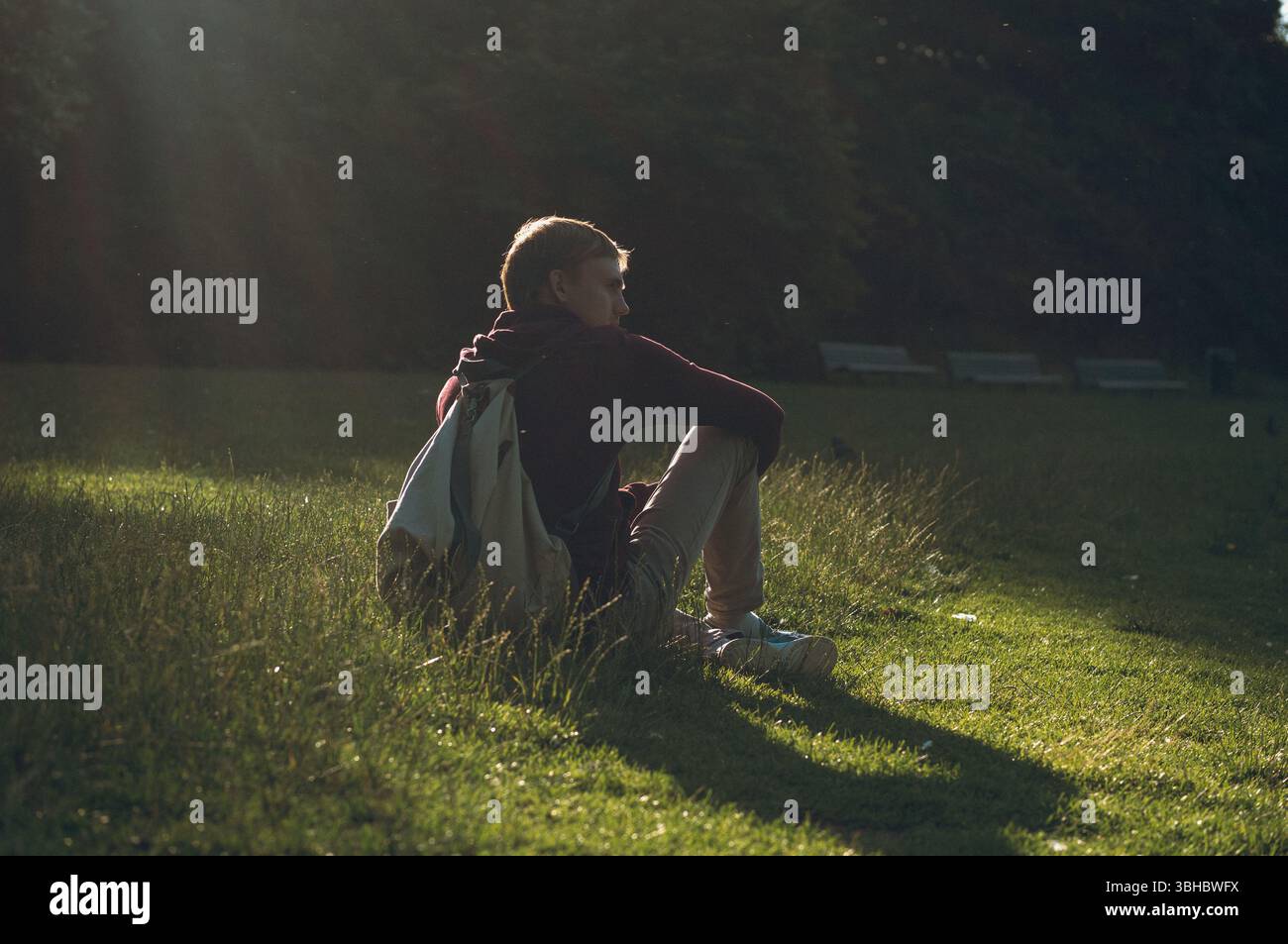 Man sitting on grass with backpack in a sunlit park Stock Photo