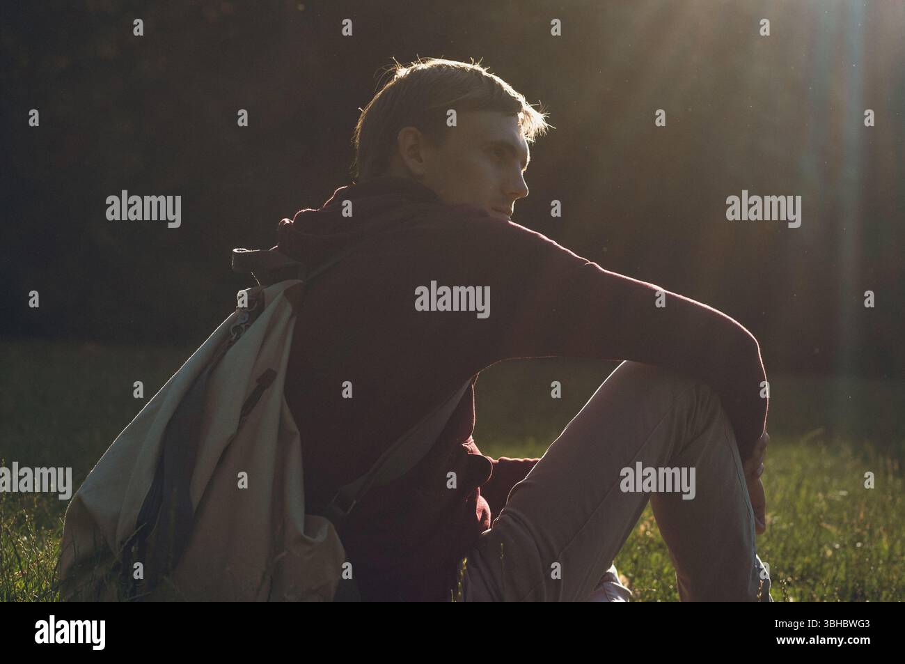 Young man relaxing on grass with backpack under warm sunlight Stock Photo