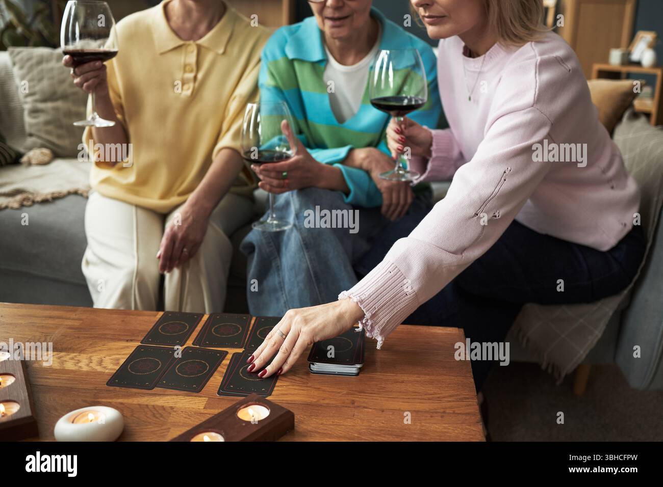 Three middle aged Caucasian women sitting together holding wine glasses, playing card game on wooden table, one woman reaching for card, candles burning in foreground Stock Photo
