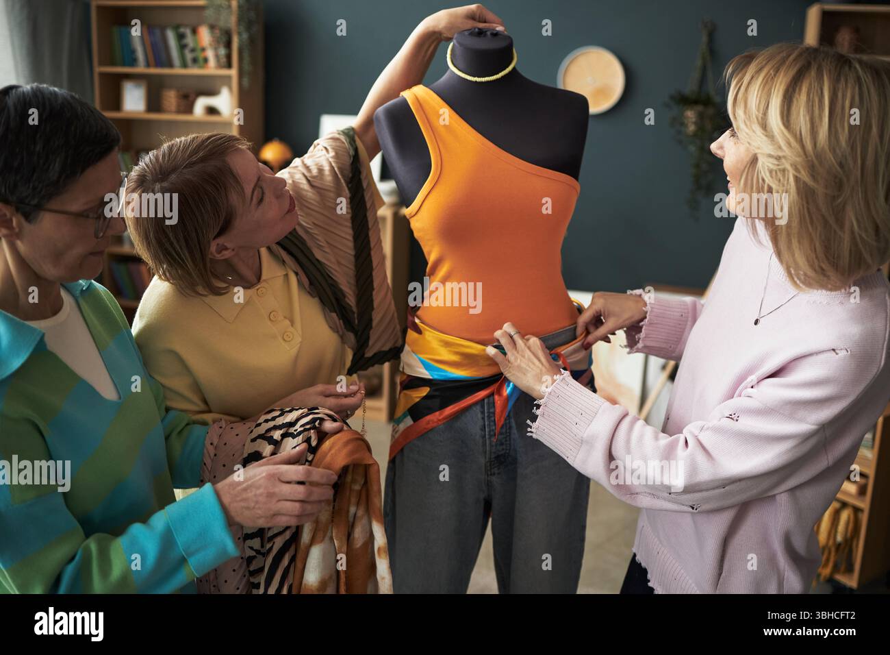 Three Caucasian middle aged women collaborating while styling mannequin with colorful top and scarf, focusing on creative teamwork and fashion arrangement in cozy indoor setting Stock Photo
