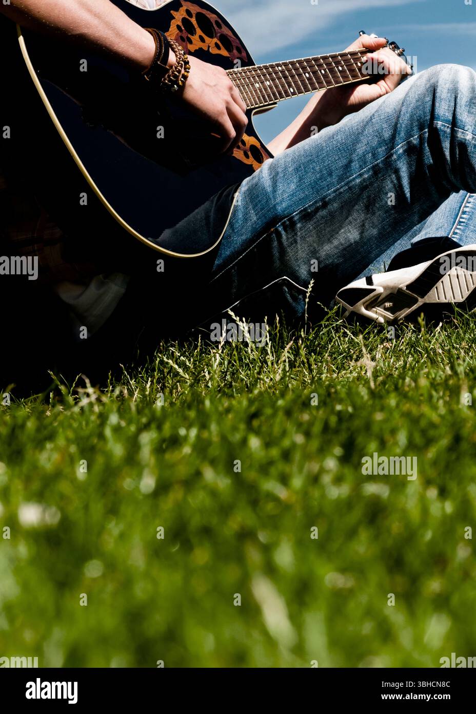 A person sits on the grass and plays a black acoustic guitar. The guitar has unique brown patterned cutouts on the body. The guitarist wears a plaid s Stock Photo