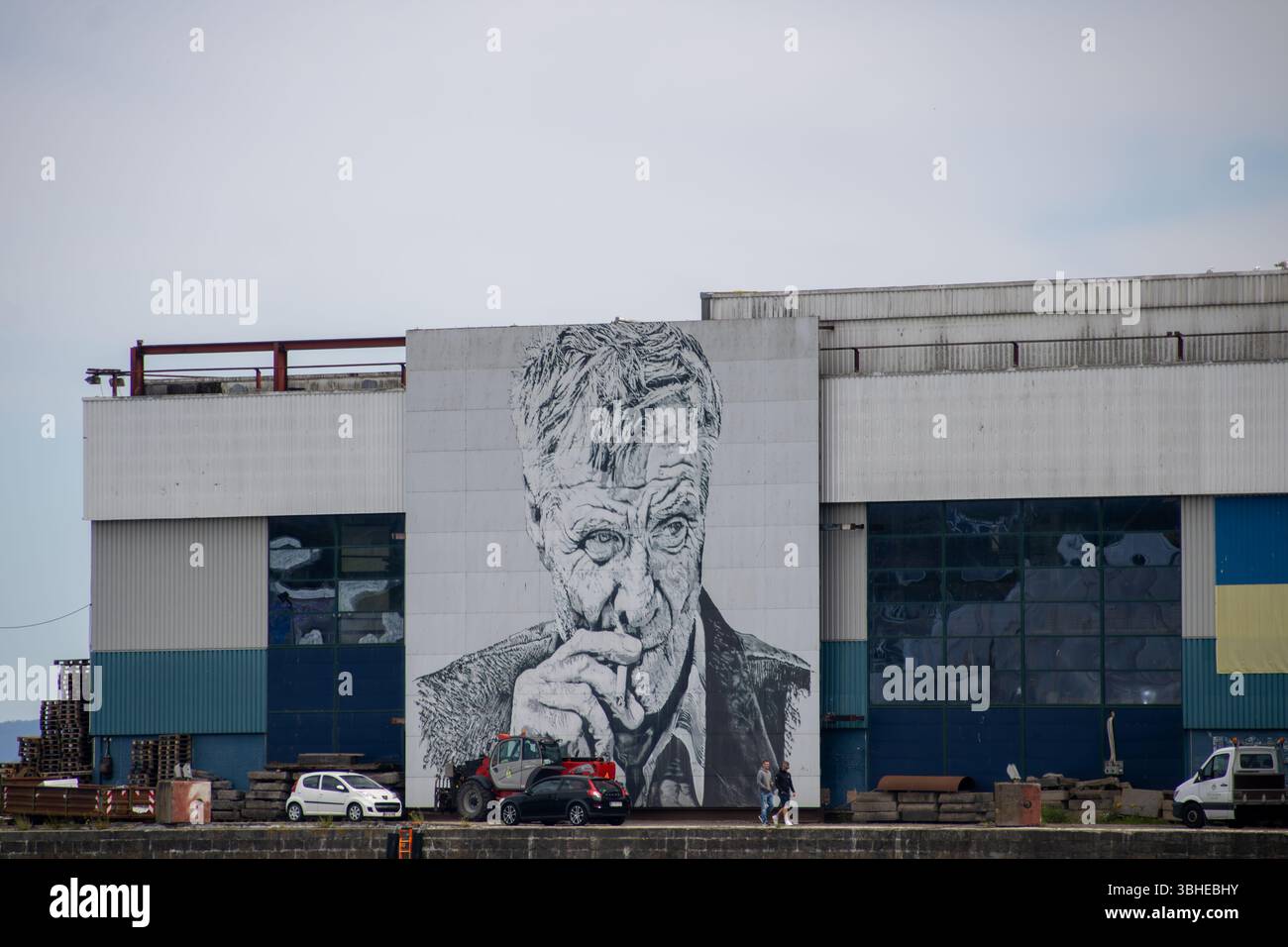 Aarhus, Dänemark: Große Schwarz‑Weiß‑Wandmalerei mit nachdenklichem älteren Mann vor Hafen‑Lagerhalle und Autos Stock Photo
