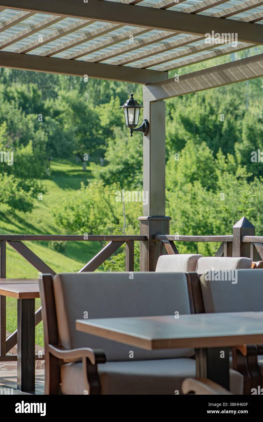 A cozy terrace with soft chairs and wooden furniture, a canopy and lantern, set against a green hill and dense forest lit by bright sunlight. Stock Photo