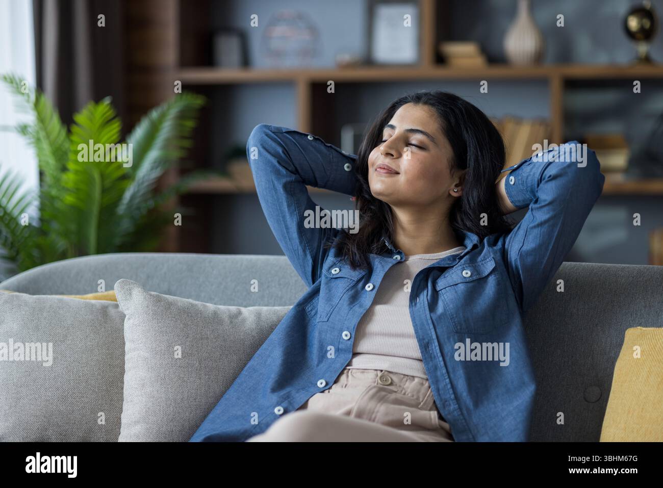 A serene young woman relaxes on a comfortable couch, hands behind her ...