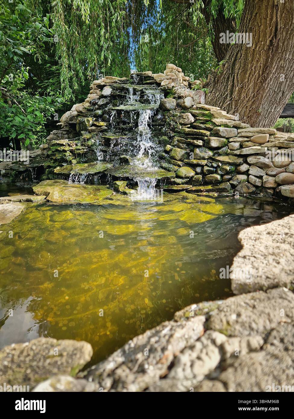 Graceful spring with cascades over moss-covered stones into a crystal-clear pond with thrown coins. Golden sunlight dances on the fountain rippling wa - Smartphone Captured Stock Image