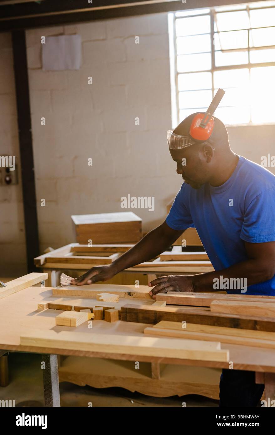 Carpenter using sanding block on wooden plank at workbench inside workshop with sunlight filtering Stock Photo