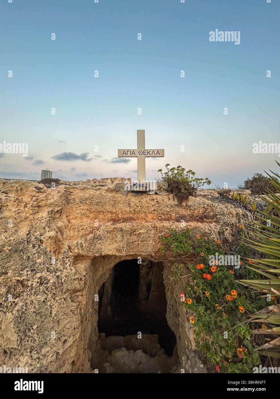 Stone entrance to an ancient cave tomb marked by a cross reading 'Saint Thecla' in Greek, with flowers and a serene sky in the background. - Smartphone Captured Stock Image