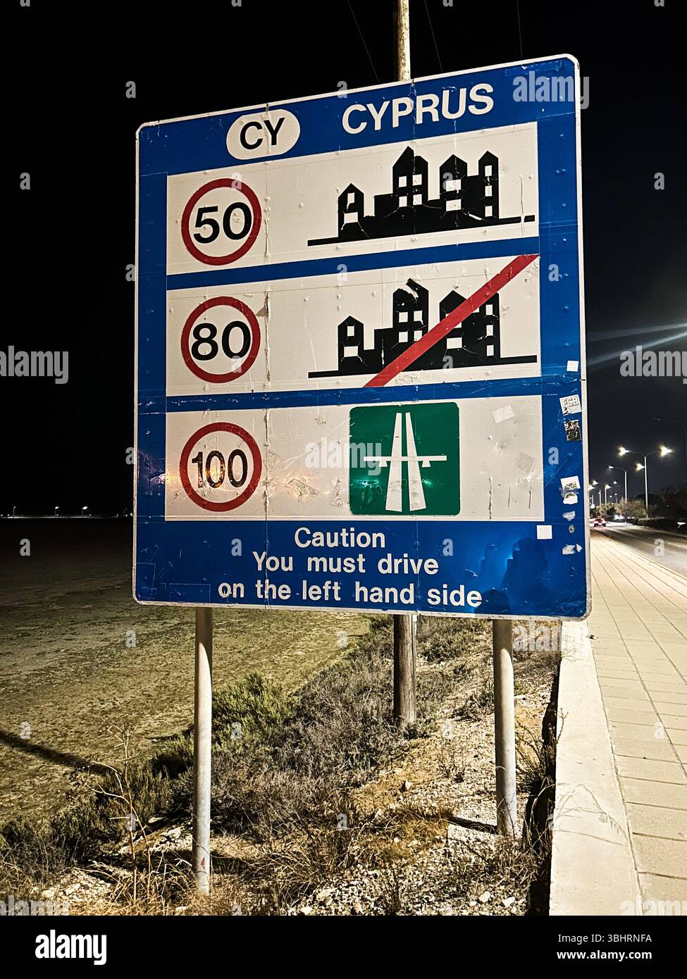 Nighttime photo of a Cyprus road sign showing speed limits for different areas and a reminder to drive on the left-hand side. Illuminated by streetlig - Smartphone Captured Stock Image