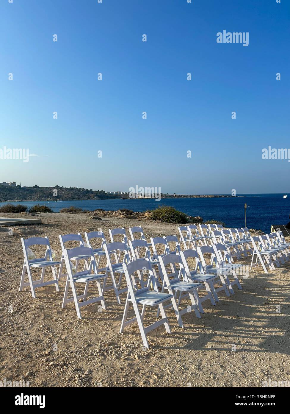 Rows of white chairs set up for an outdoor wedding ceremony on a rocky coast, overlooking the deep blue sea under a clear sky. Perfect for event, cele - Smartphone Captured Stock Image