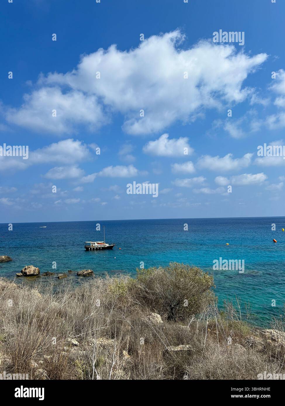 A small boat floats peacefully on turquoise Mediterranean waters under a bright blue sky with scattered clouds, near a rocky, dry coastline. - Smartphone Captured Stock Image