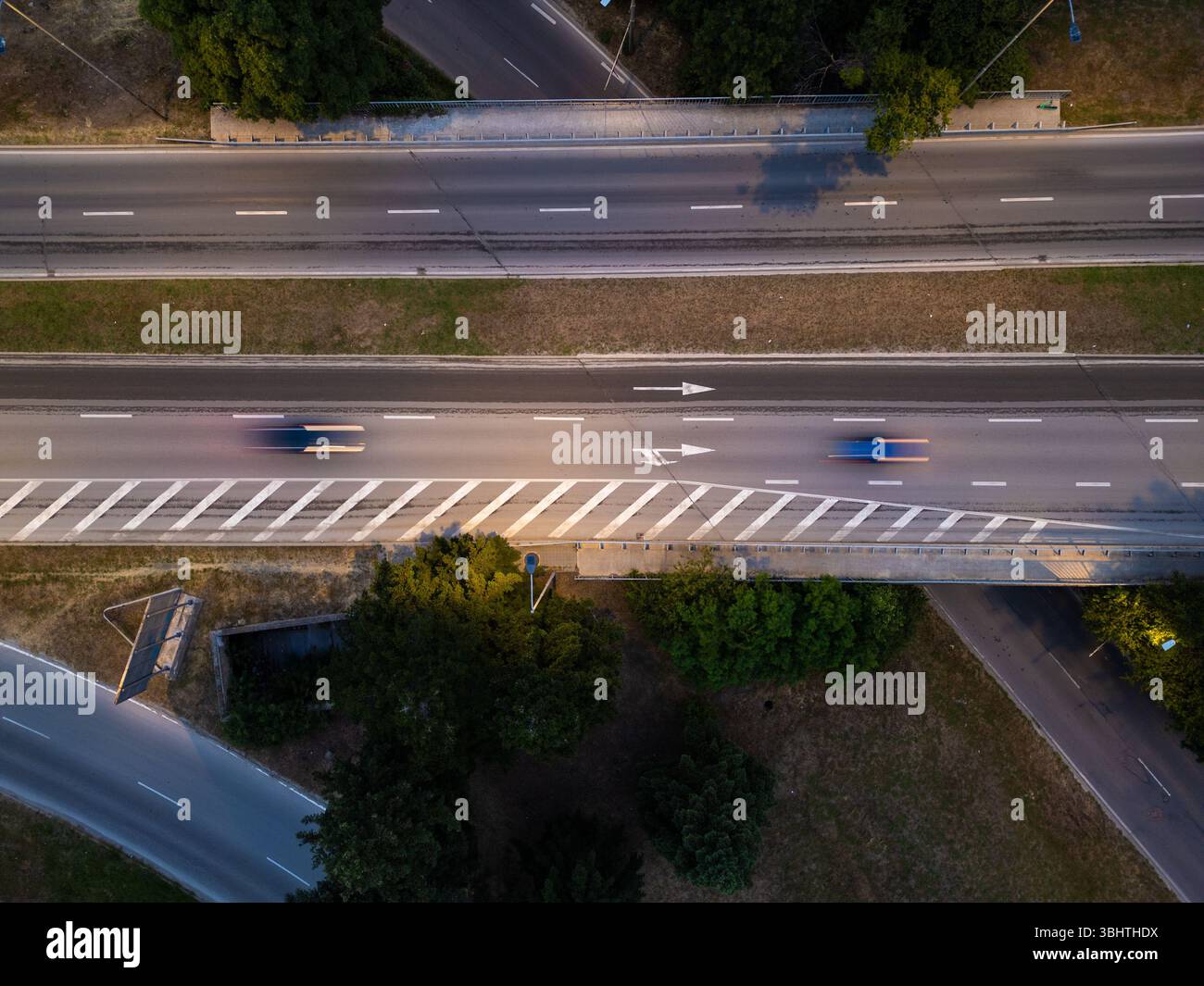 Aerial view of multilane highway with fast-moving cars at twilight. Motion blur emphasizes speed. Urban infrastructure with directional arrows, trees Stock Photo