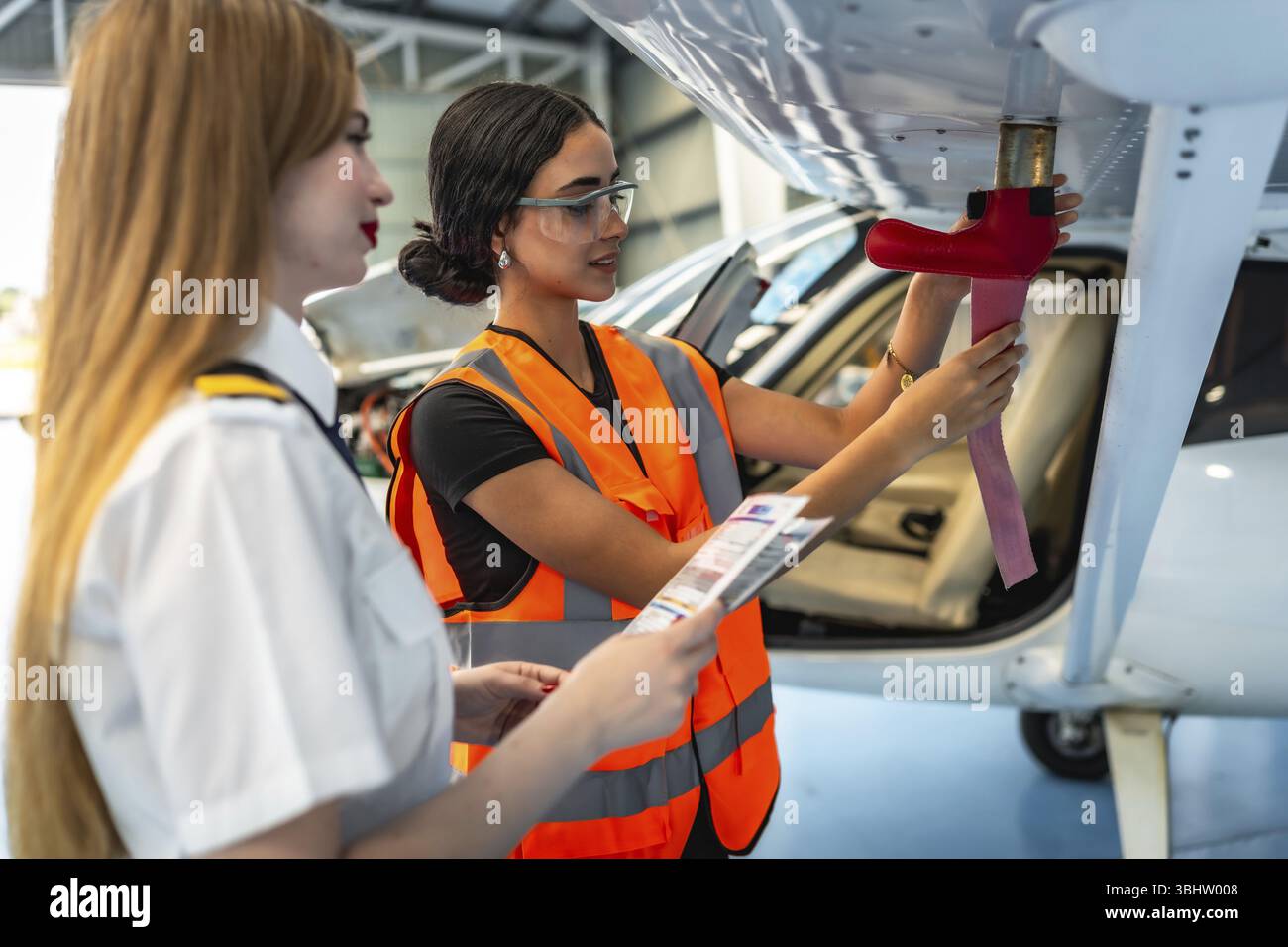 Female aircraft maintenance engineer inspecting airplane wing with pilot assisting, holding checklist and discussing procedures in hangar Stock Photo