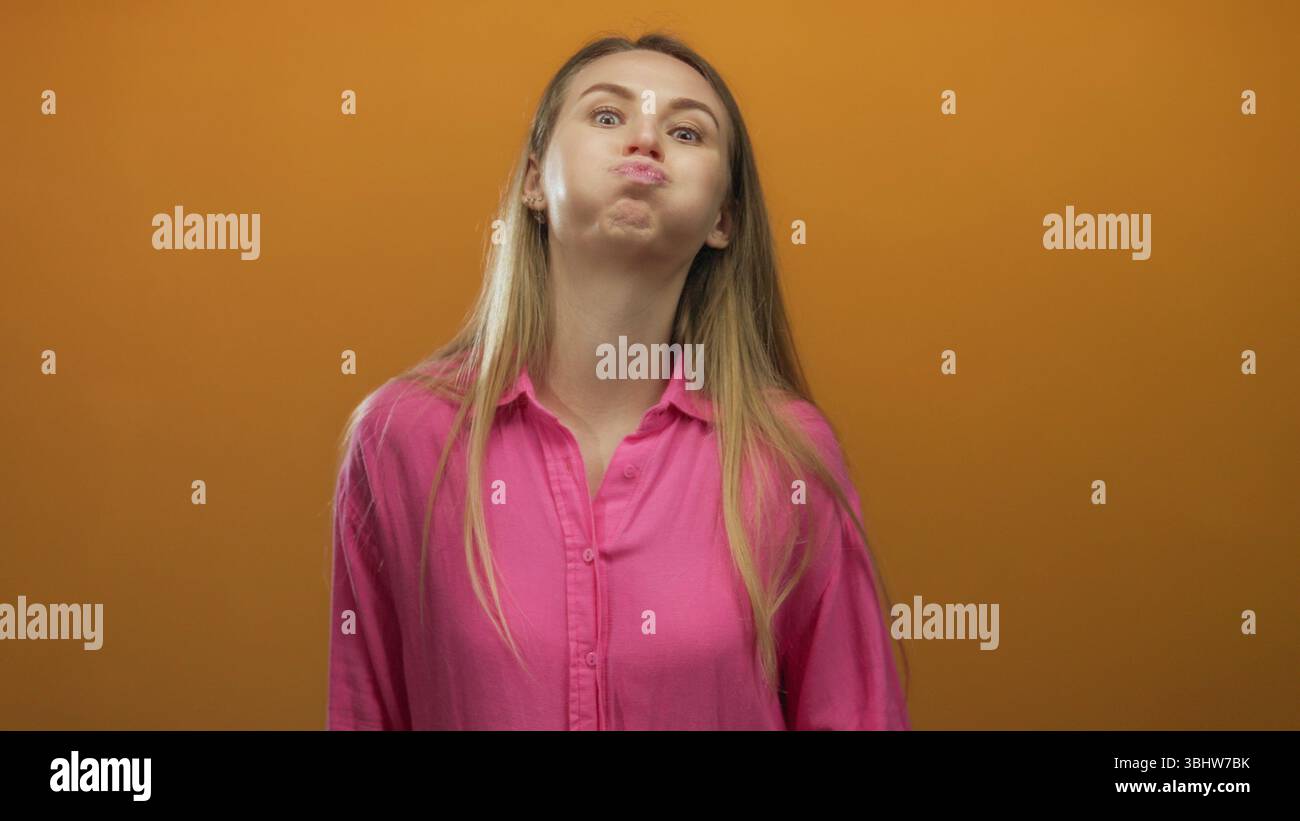 Woman with long hair and pink shirt playfully puffing cheeks against an isolated orange background, expressing silly emotions with a joyful expression Stock Photo