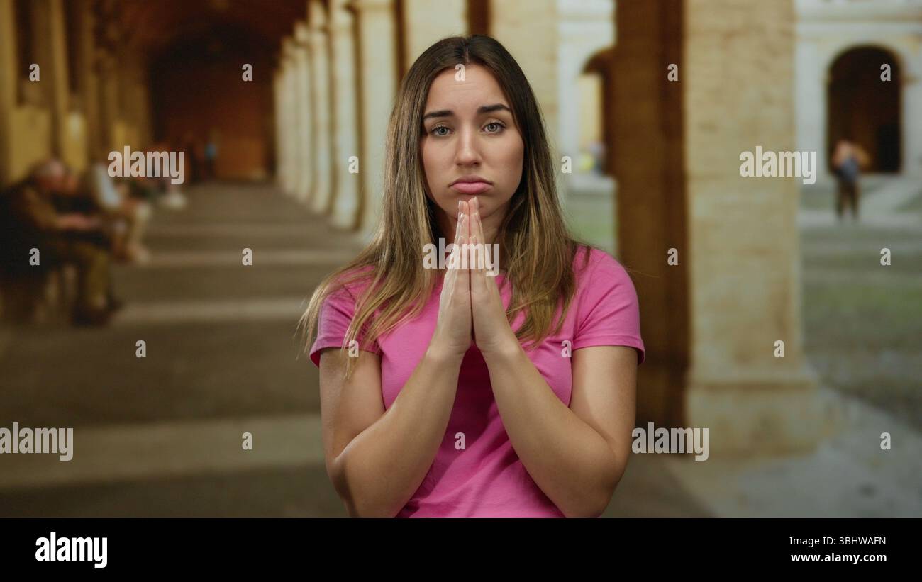 Woman in pink shirt praying with eyes closed in ancient university courtyard. Stock Photo