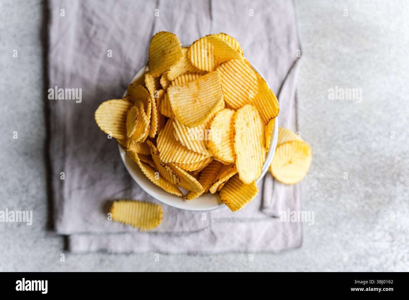 A bowl filled with delicious homemade potato chips, showcasing a golden, crispy texture, resting on a soft gray cloth, perfect for a snack or a casual Stock Photo